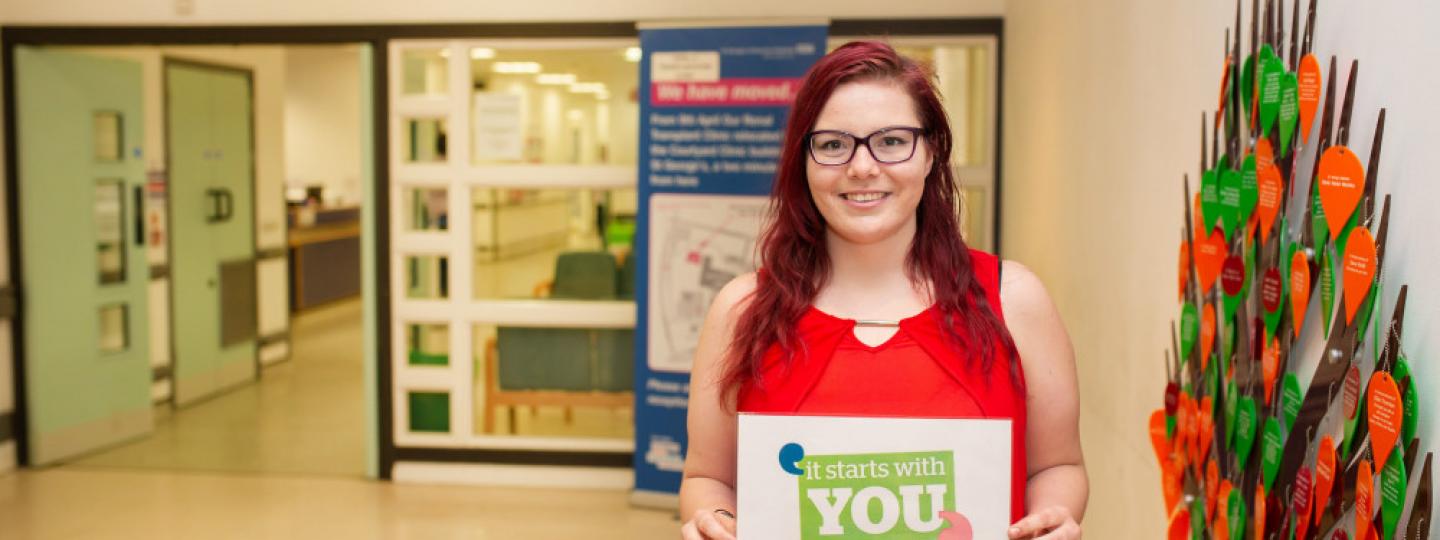 Image of young woman standing in the hall of a hospital holding sign It starts with you